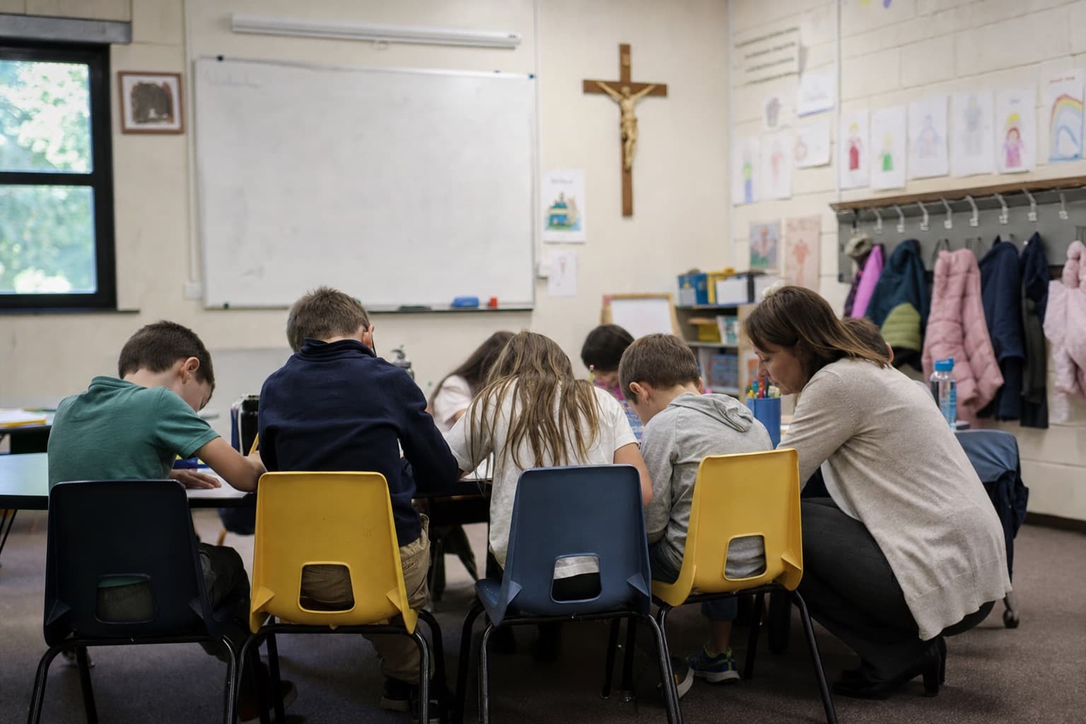 A catechist kneeling beside children at their desks in a parish classroom